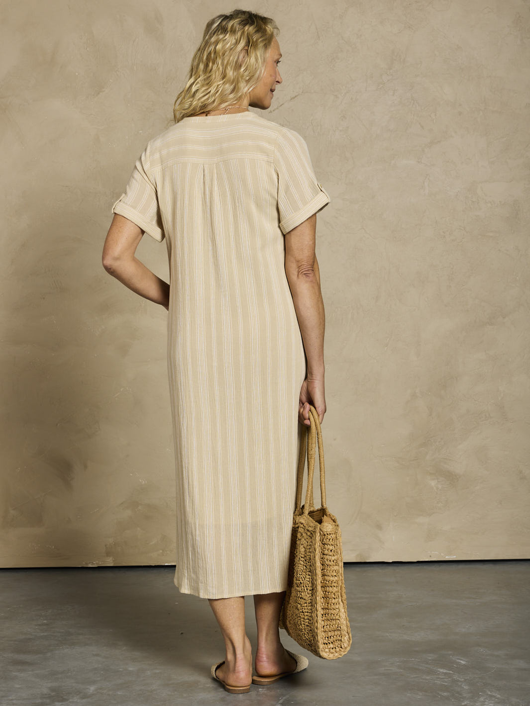 Woman wearing a beige dress holding a woven bag against a plain background