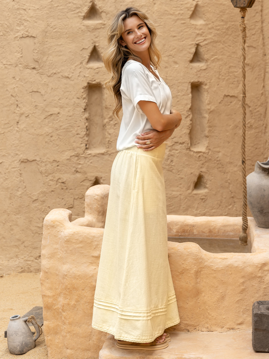 Woman in a white top and light yellow skirt standing in front of a textured beige wall.