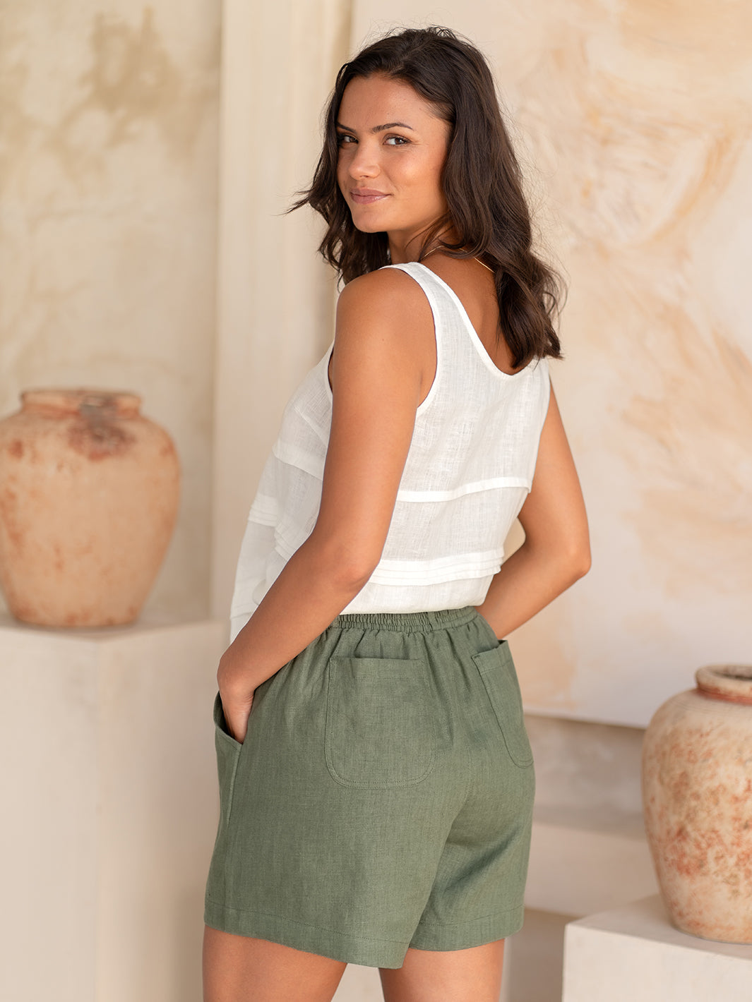 Woman wearing a white sleeveless top and green shorts against a neutral background with vases.