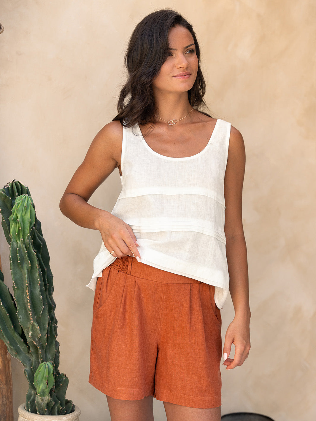 Woman wearing a white sleeveless top and orange shorts standing against a beige wall with a cactus plant.
