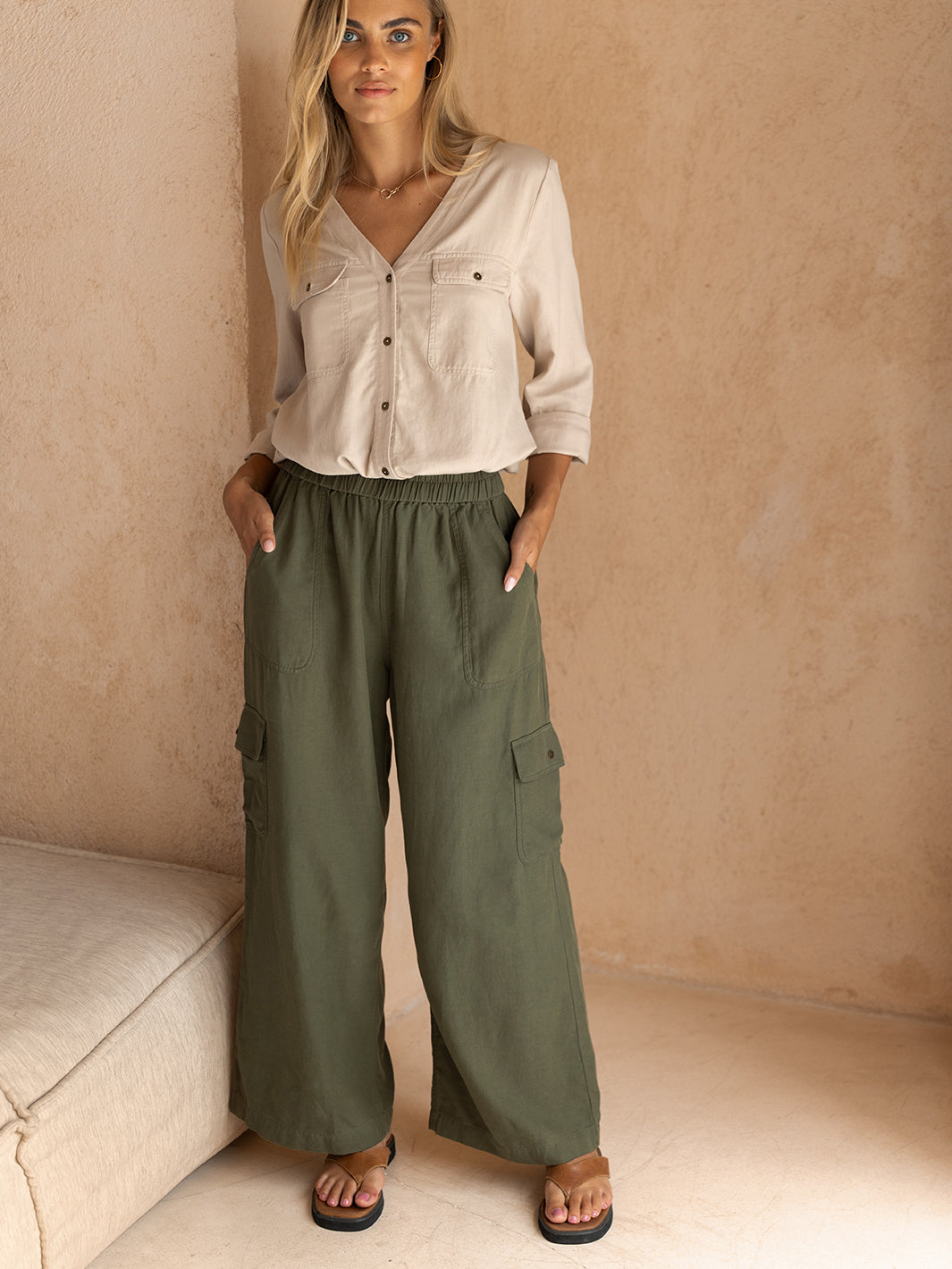 Woman wearing a beige shirt and green cargo pants against a beige wall.