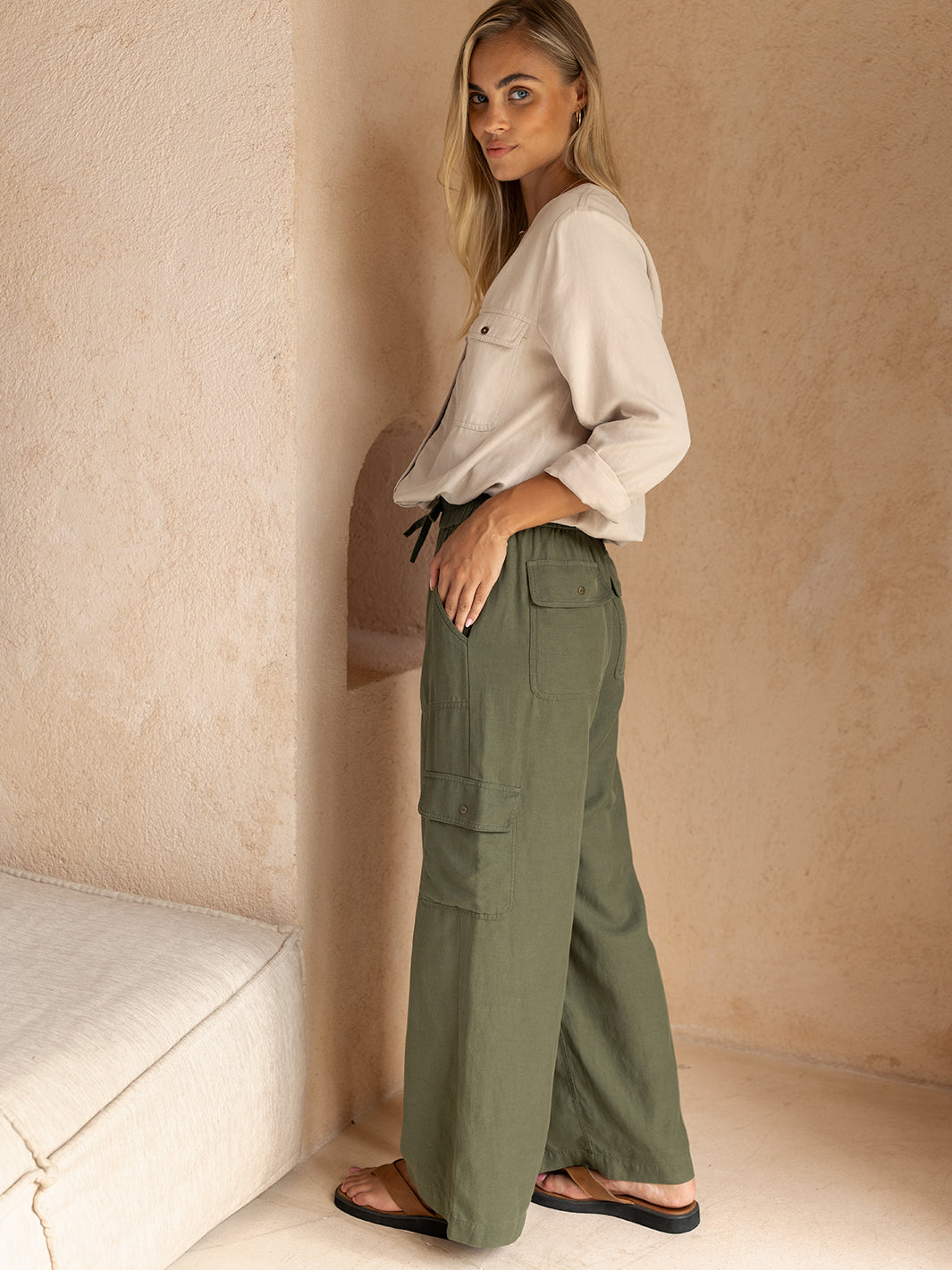 Woman wearing a white blouse and green cargo pants against a beige wall.