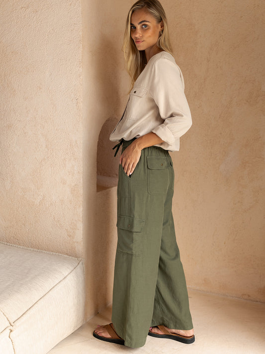 Woman wearing a white blouse and green cargo pants against a beige wall.
