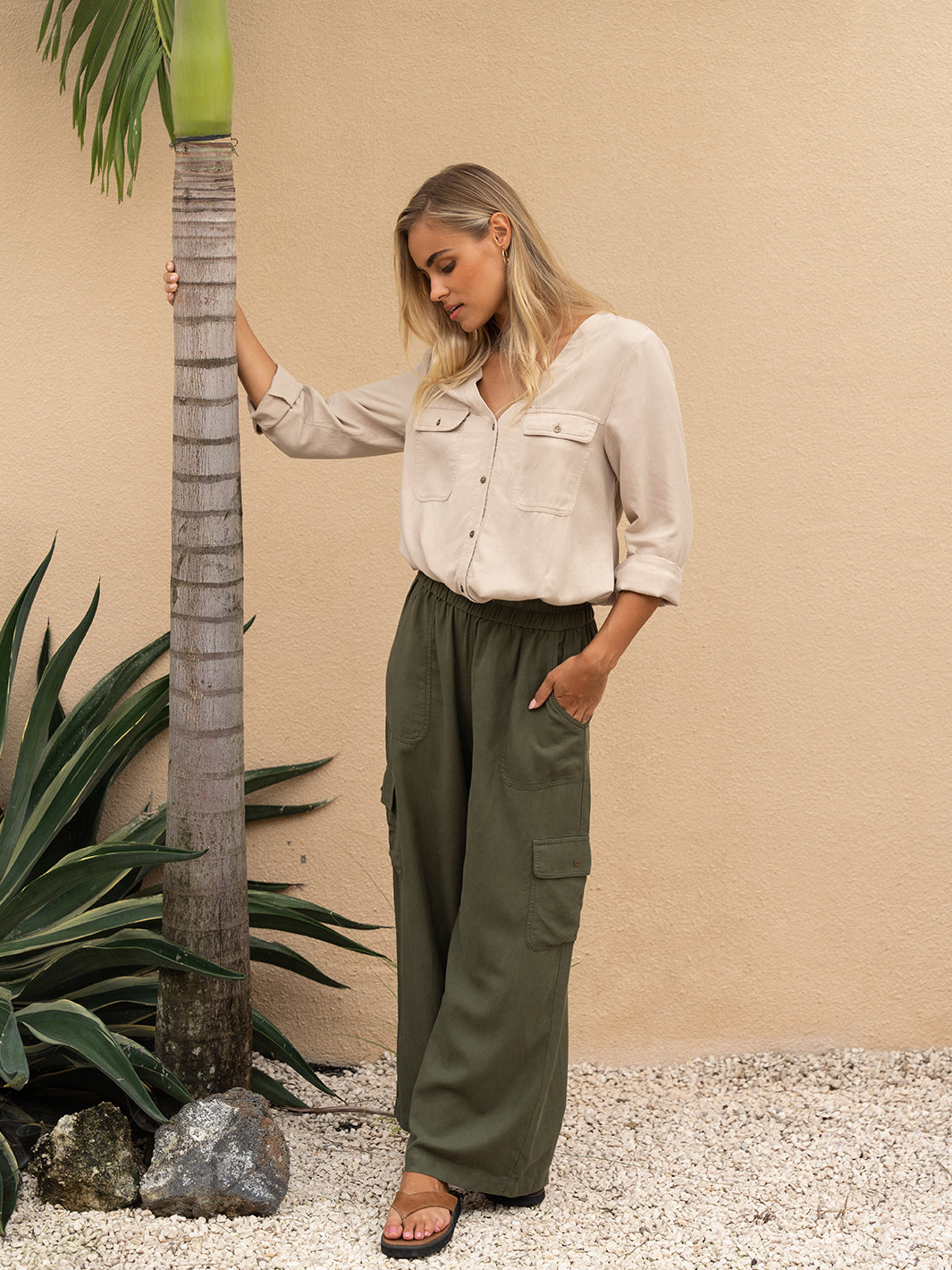 Woman in beige shirt and green pants standing next to a palm tree against a beige wall.