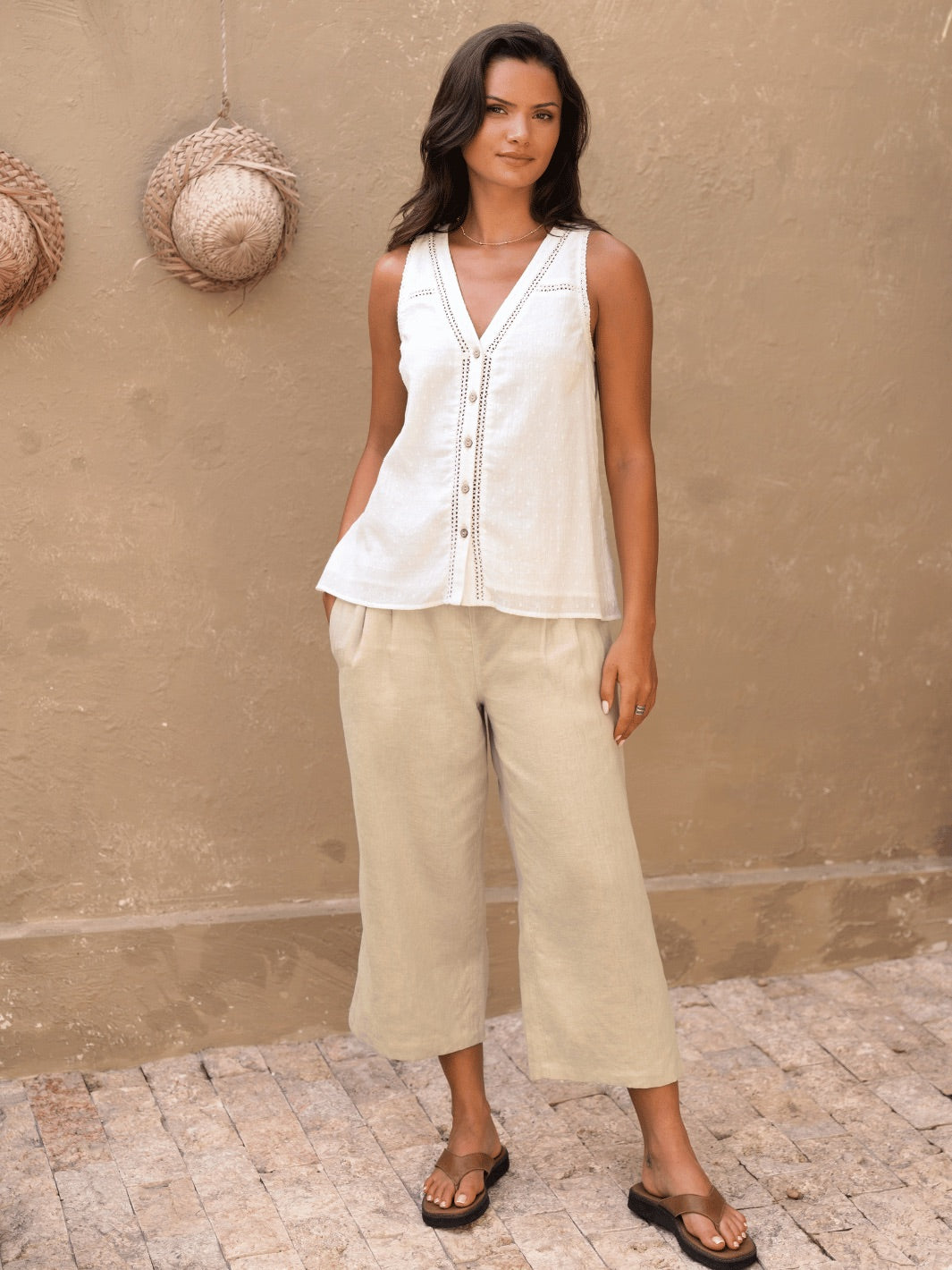 Woman wearing a white sleeveless top and beige pants against a beige wall.