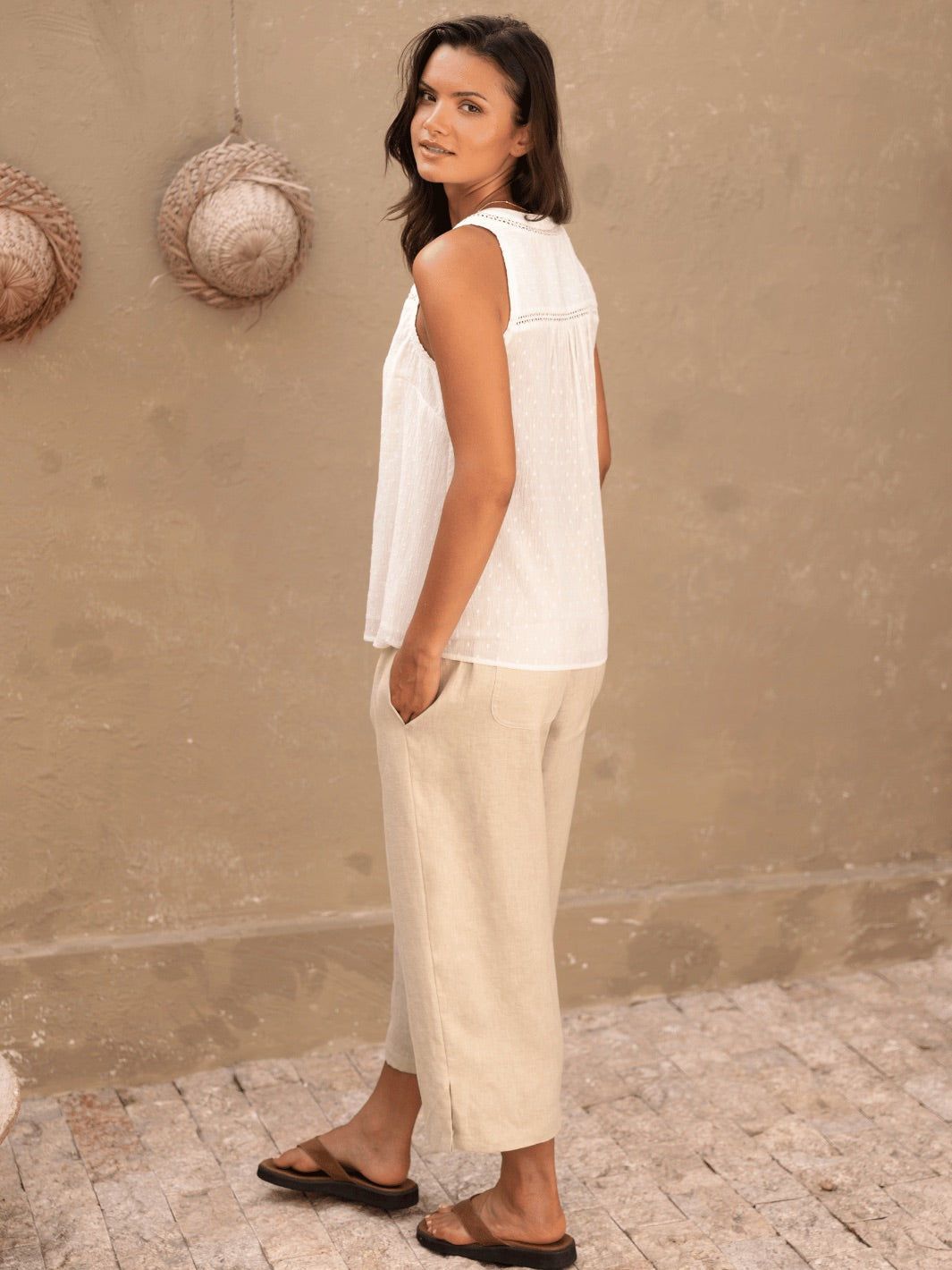 Woman wearing a white sleeveless top and beige pants standing against a beige wall.