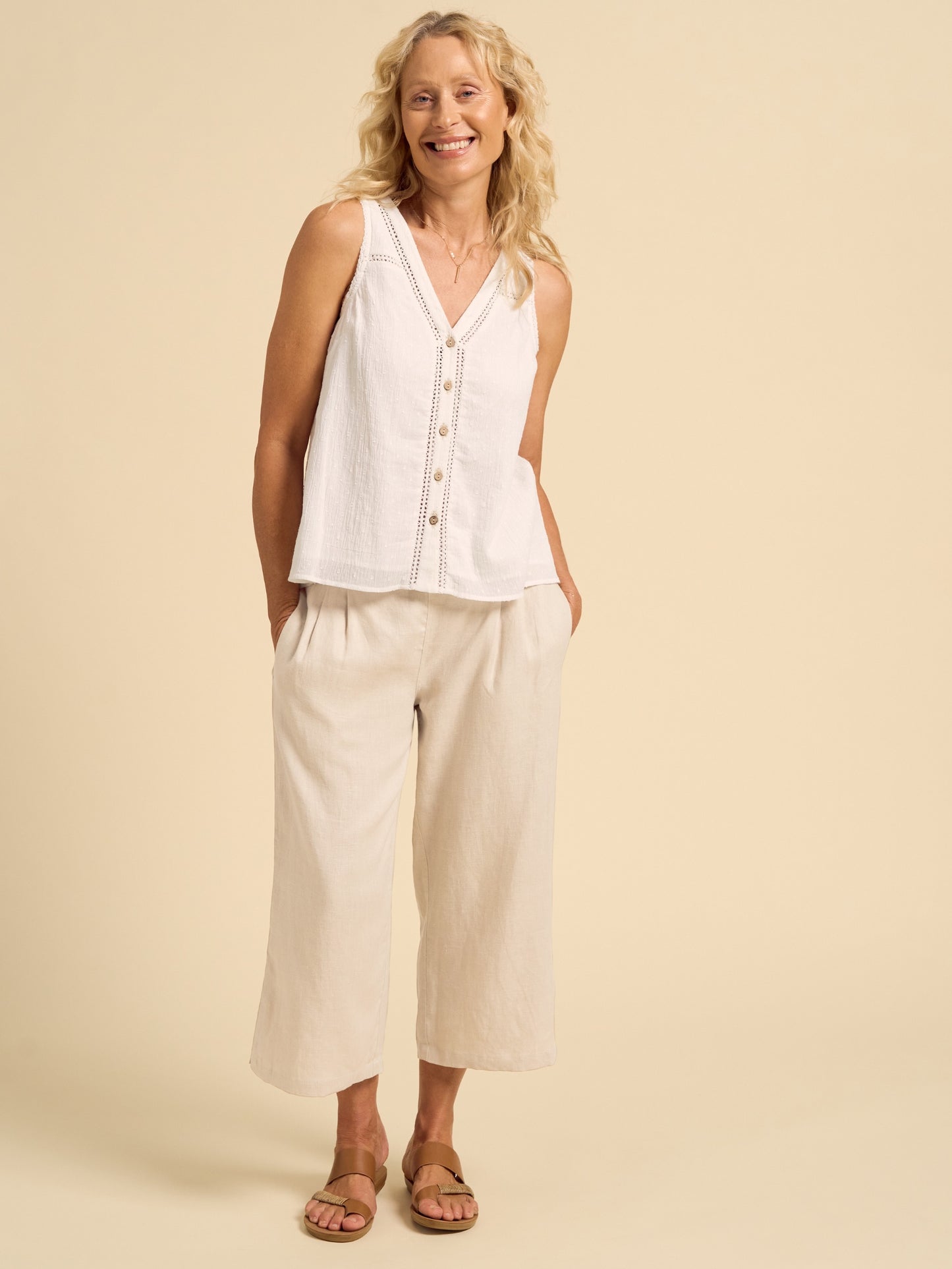 Woman wearing a white sleeveless top and beige pants against a beige background