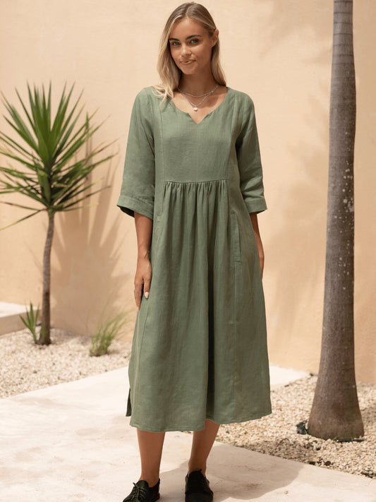 Woman wearing a green dress standing in a desert-like setting with plants and a beige wall.