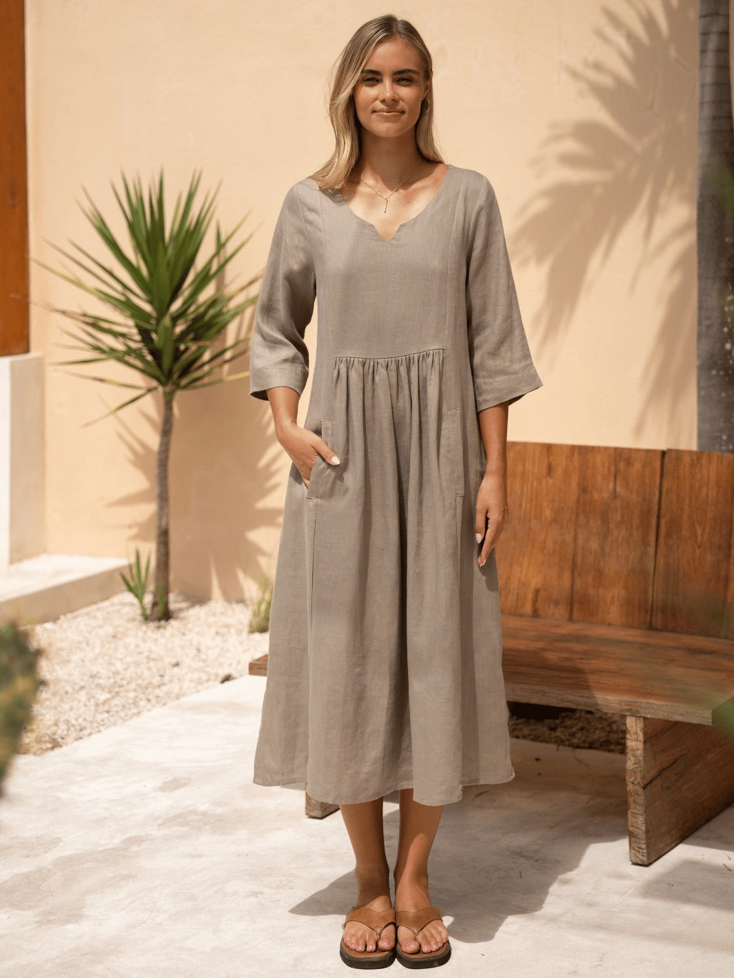 Woman wearing a long, gray dress standing outdoors with a wooden bench and plant in the background.