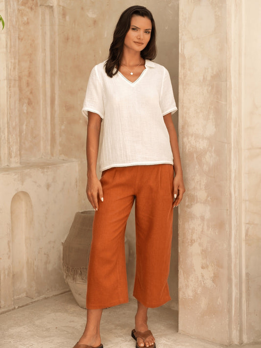 Woman wearing a white top and rust-colored pants standing against a beige wall.