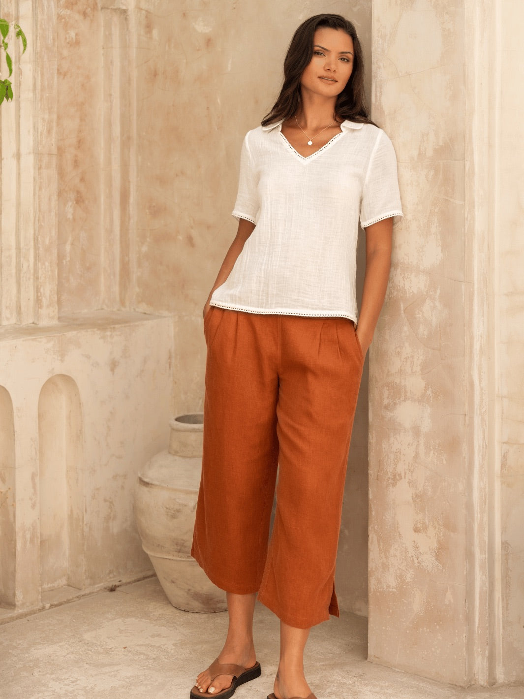 Woman wearing a white top and rust-colored pants standing against a beige wall.