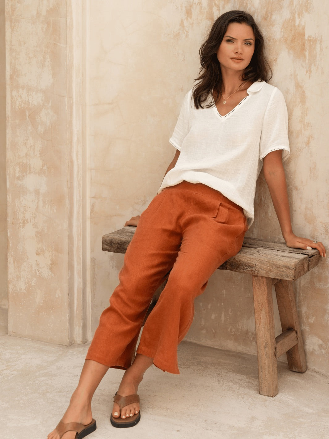 Woman sitting on a wooden bench wearing a white top and rust-colored pants against a beige wall.