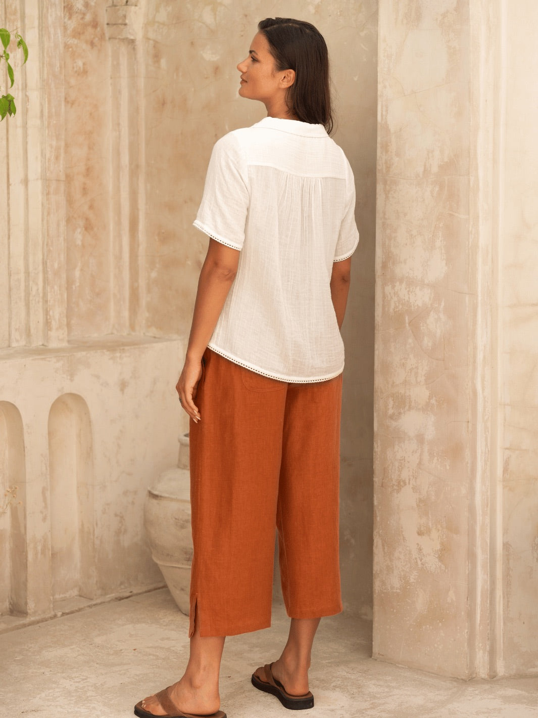 Woman wearing a white shirt and rust-colored pants standing against a beige wall.