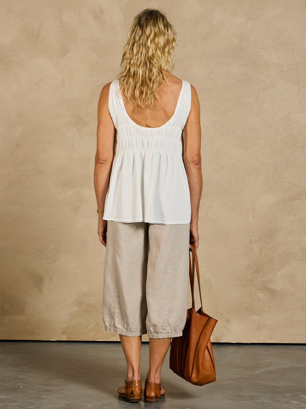 Woman wearing a white sleeveless top and beige pants, holding a brown bag against a beige wall.