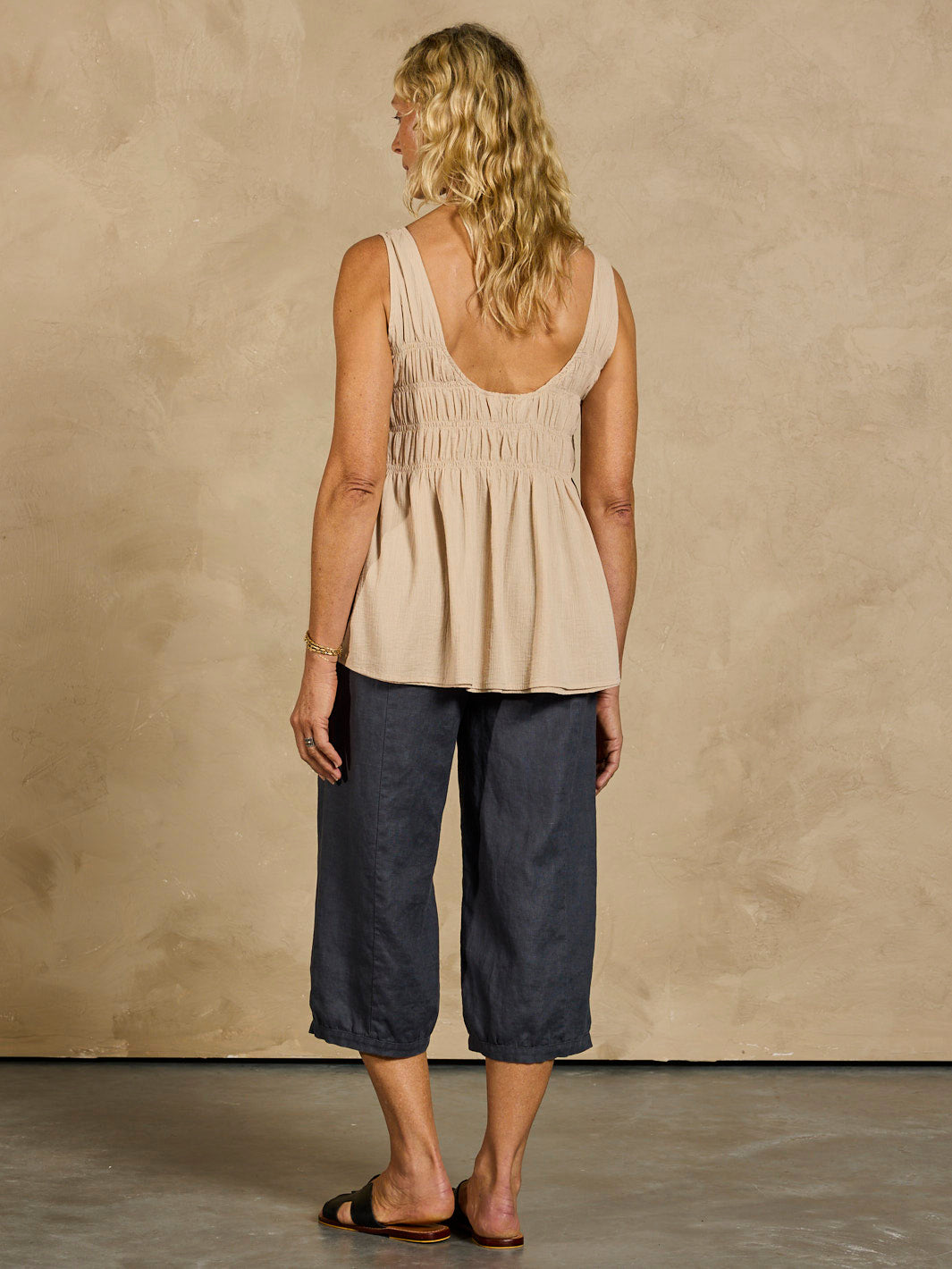 Woman wearing a beige sleeveless top and dark blue pants against a beige wall.