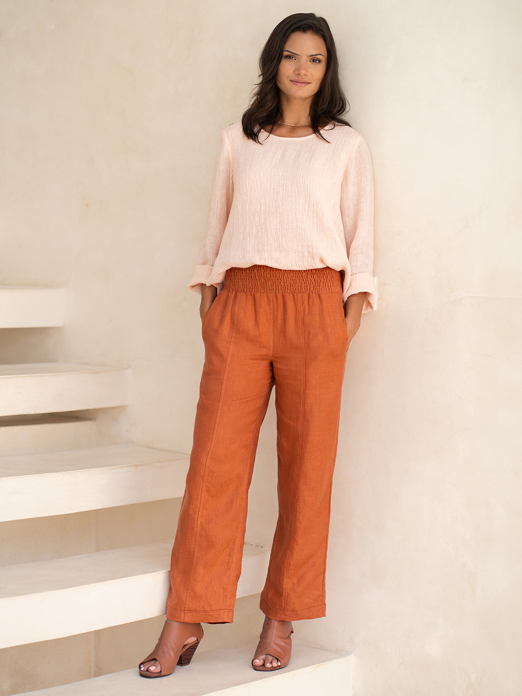 Woman wearing a light pink blouse and rust-colored pants standing on a staircase.