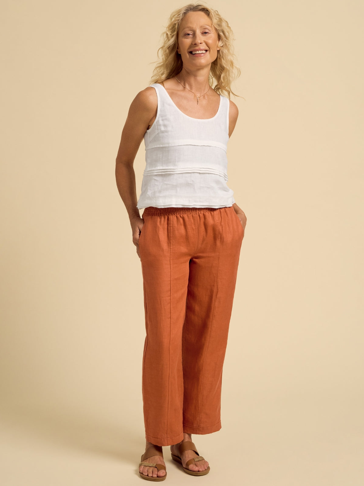 Woman wearing a white tank top and rust-colored pants on a beige background