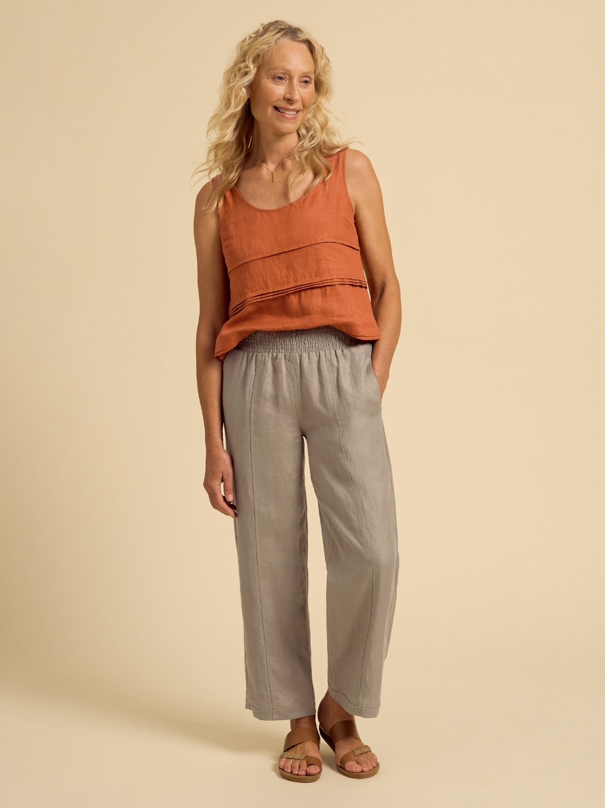 Woman wearing an orange sleeveless top and gray pants on a beige background