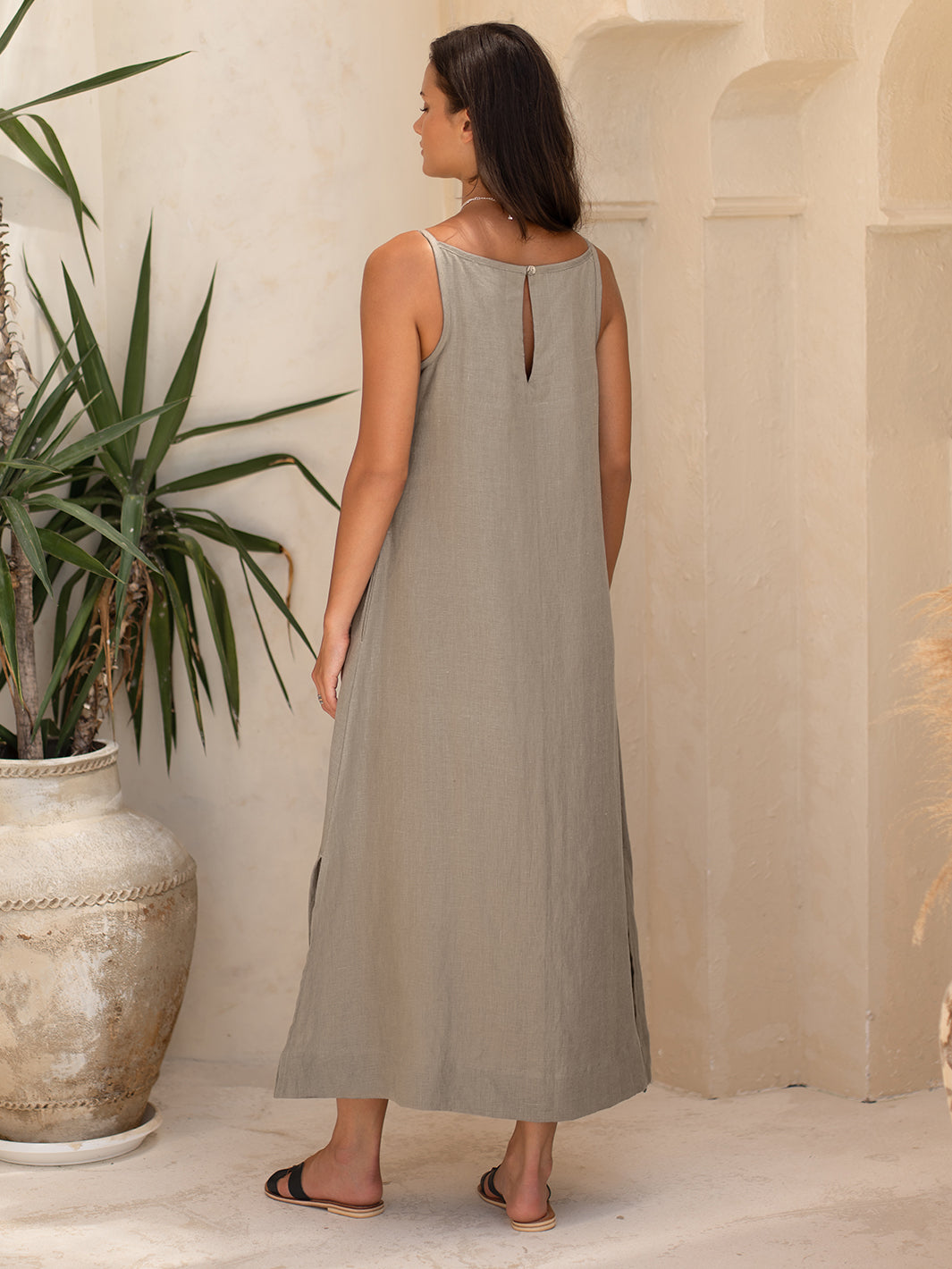 Woman wearing a long, light-colored dress standing in a sunlit room with a plant and architectural elements.