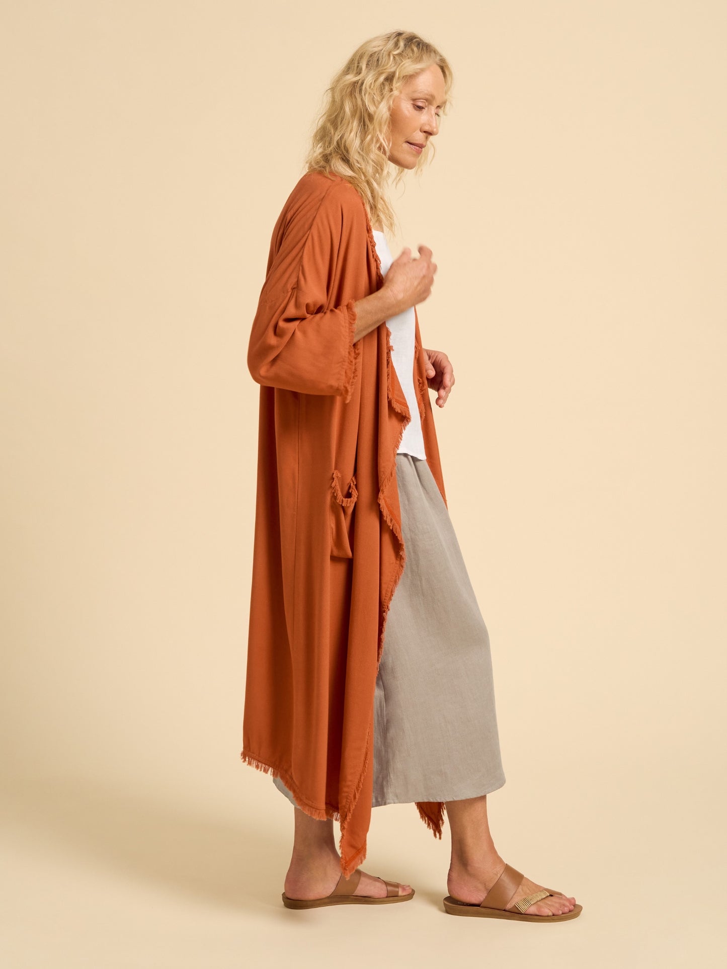 Woman wearing a long rust-colored cardigan over a white top and gray skirt on a beige background