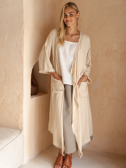 Woman wearing a long beige cardigan over a white top and gray pants, standing against a beige wall.