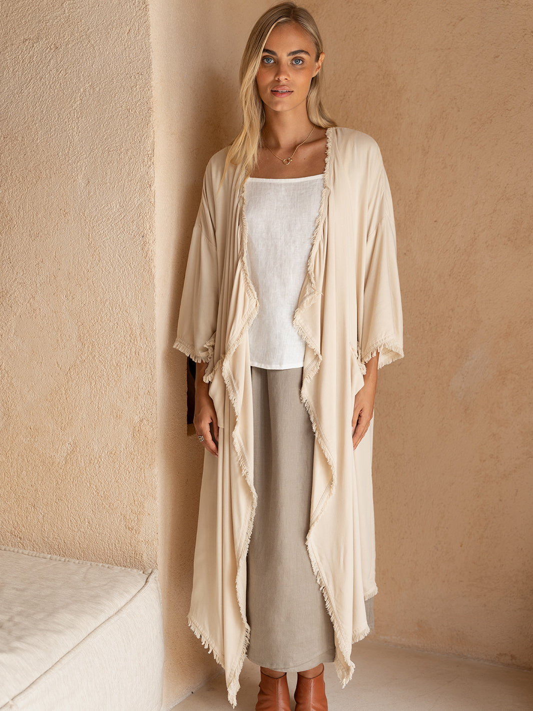 Woman wearing a beige long cardigan over a white top and gray pants against a beige wall.
