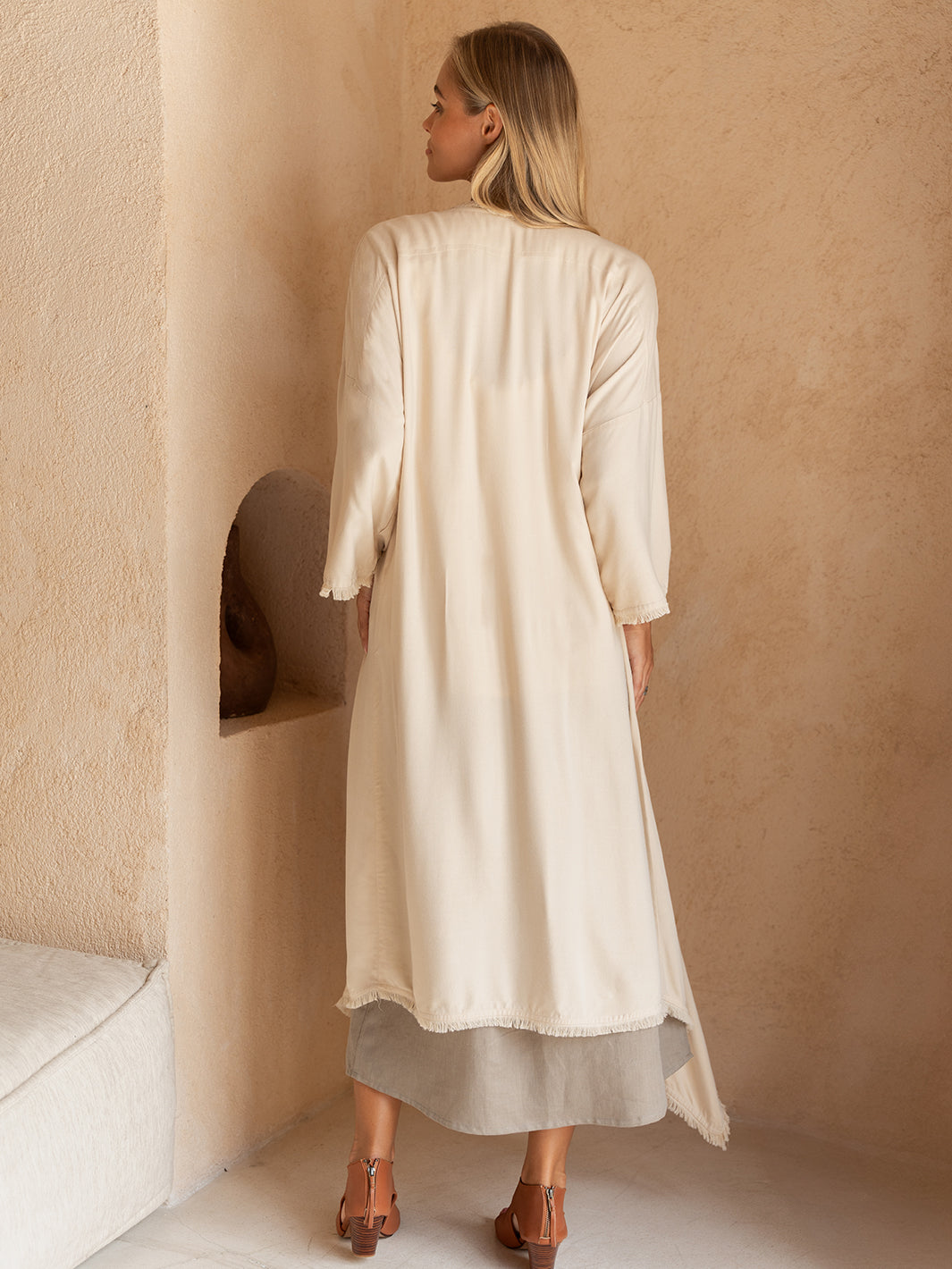 Woman wearing a long, light-colored dress standing against a beige wall.