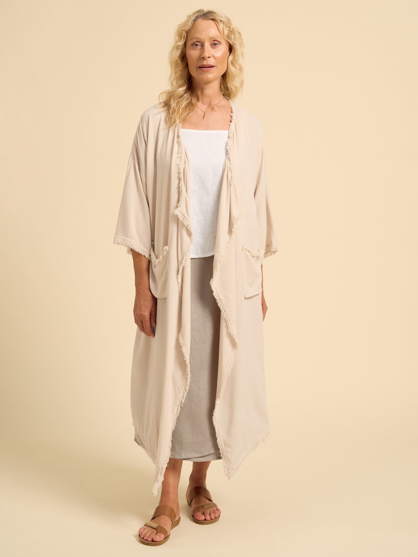 Woman wearing a beige kimono-style garment over a white top and gray pants on a beige background