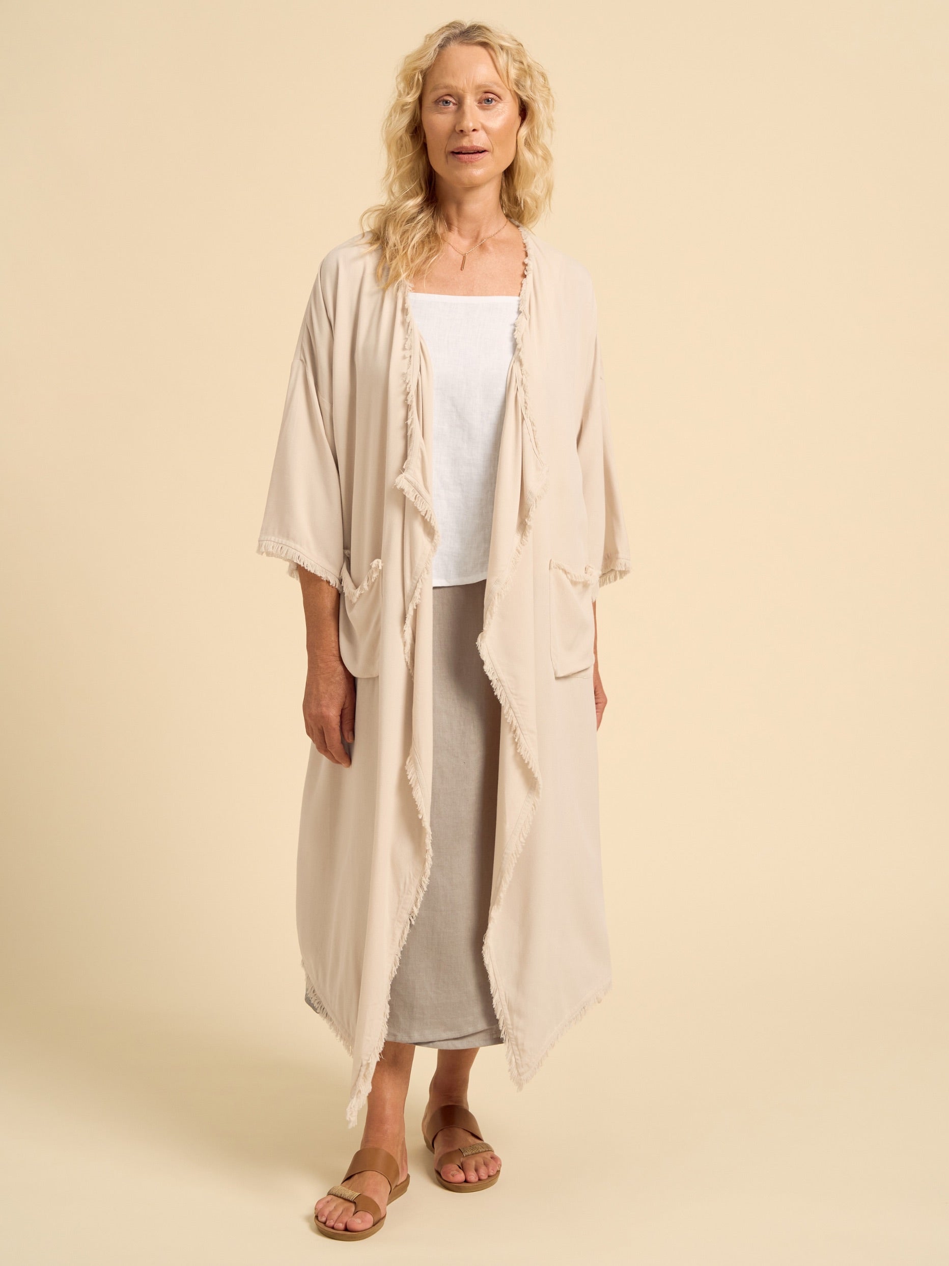 Woman wearing a beige kimono-style garment over a white top and gray pants on a beige background